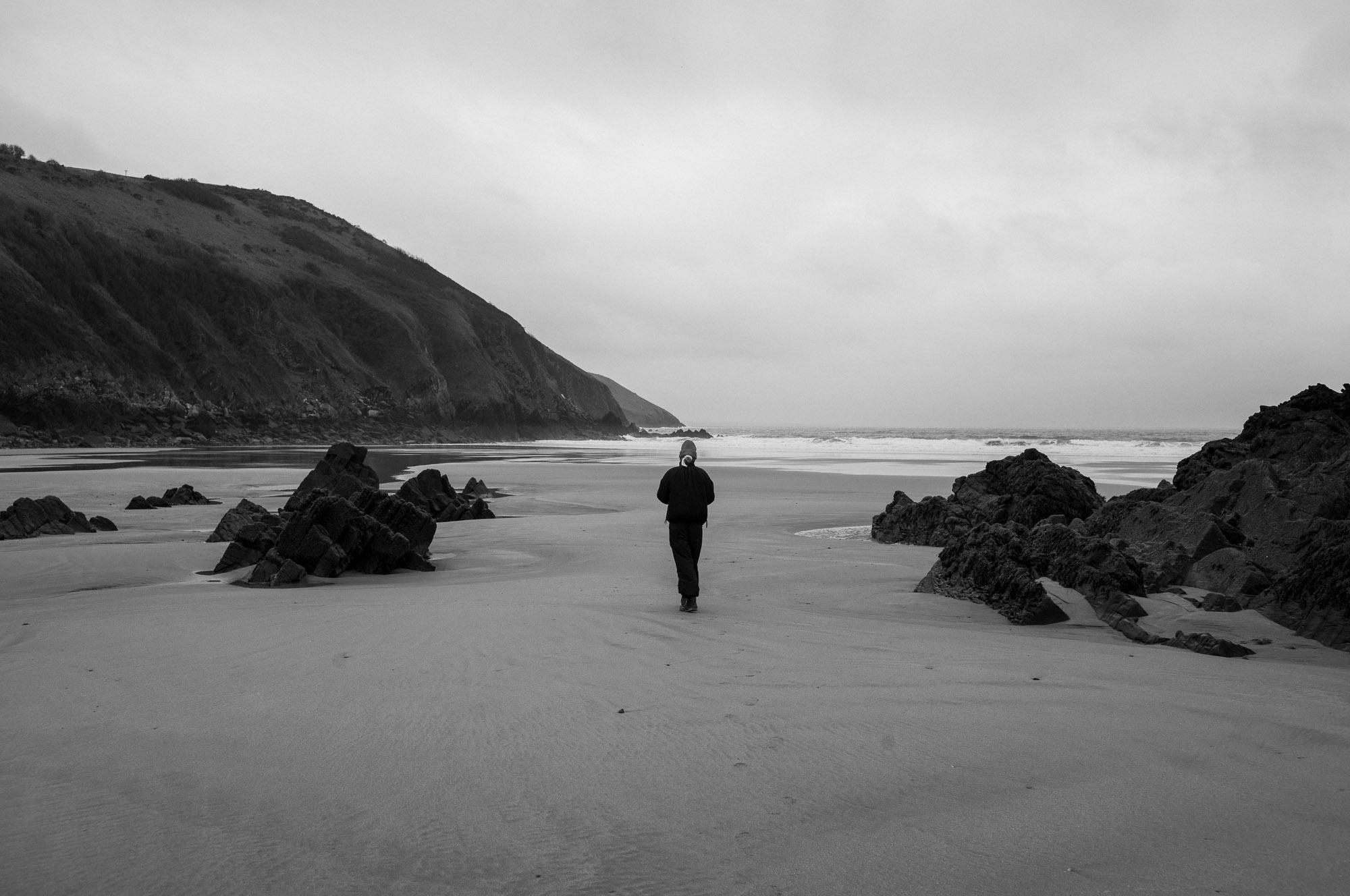 A black-and-white photograph of a lone figure walking across an empty beach toward the sea, with a headland to the left and dark rocks framing the foreground.